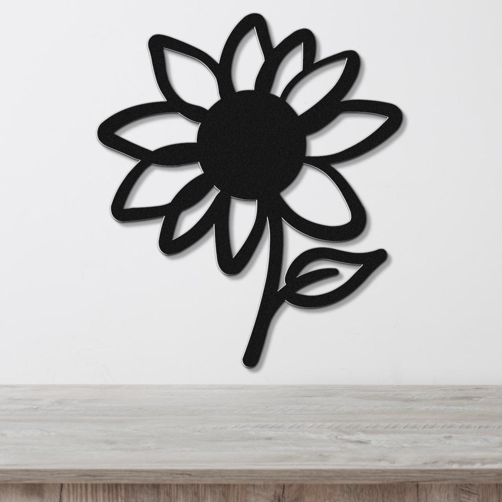 Black metal sunflower wall art with cutout petals and a leaf, mounted on a white wall above a light wood surface.