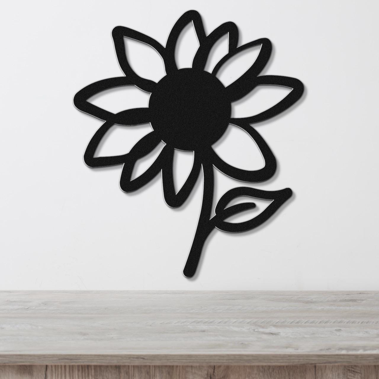 Black metal sunflower wall art with cutout petals and a leaf, mounted on a white wall above a light wood surface.