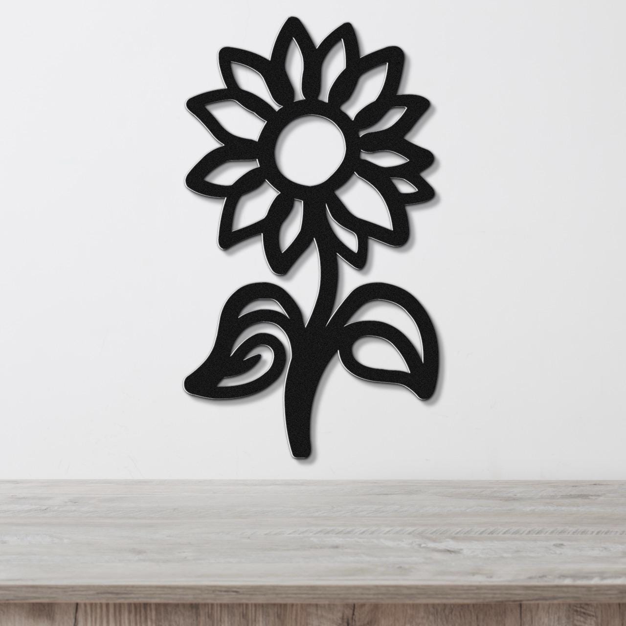 Black metal sunflower wall art with a cutout silhouette design, mounted on a white wall above a wooden surface.