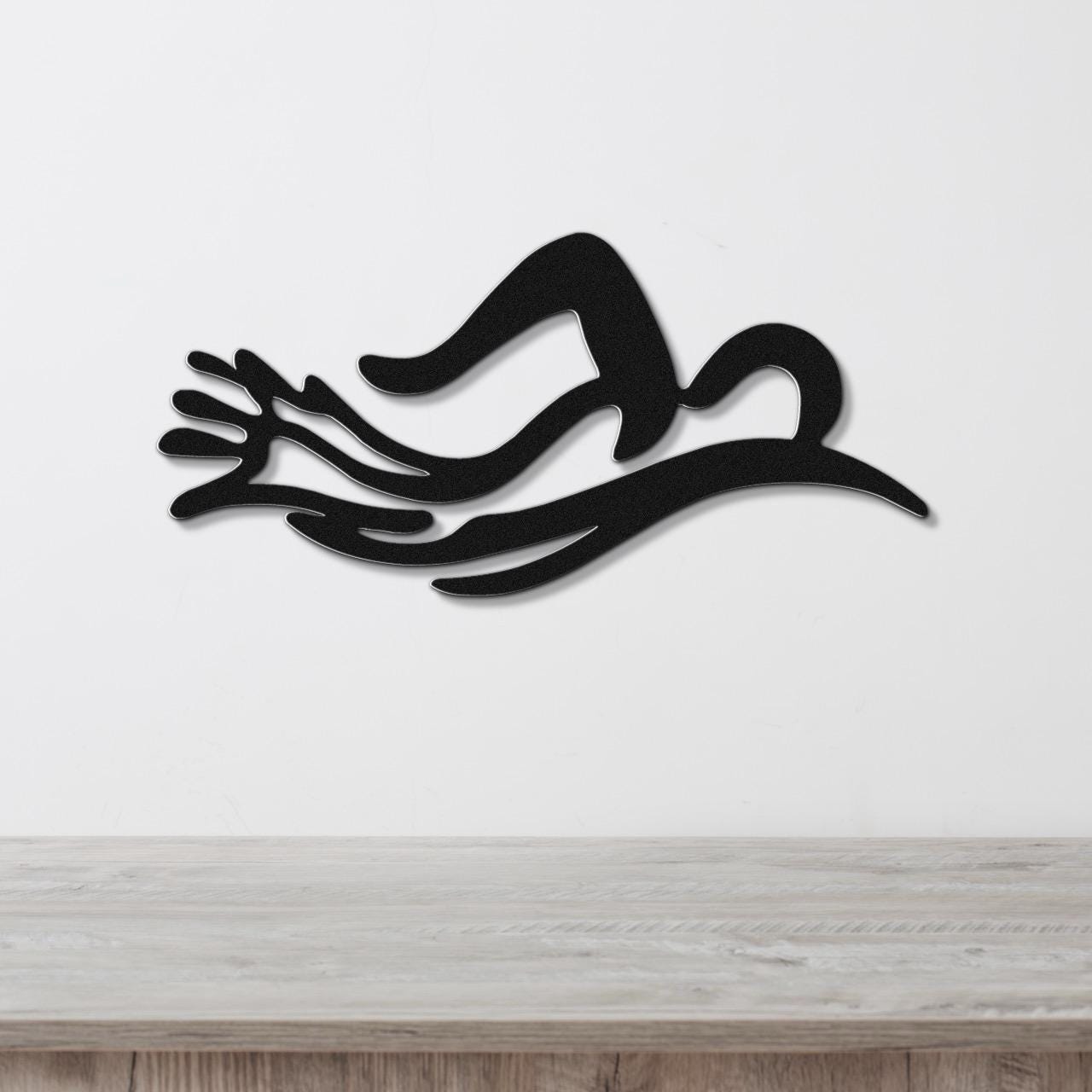 Black metal wall art in a swimmer silhouette with flowing motion lines, mounted on a white wall above a light wood surface.