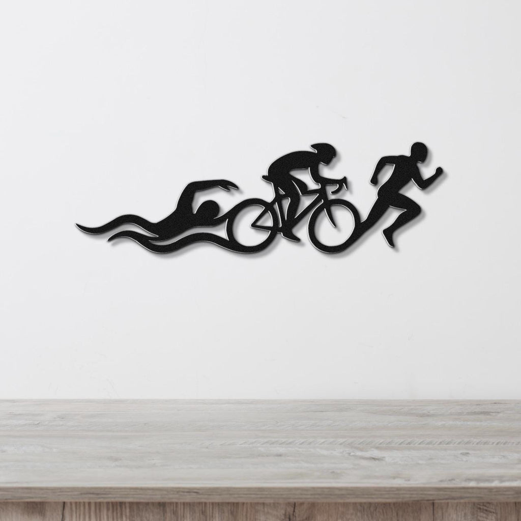 Black metal triathlon wall art with swimmer, cyclist, and runner silhouettes and motion lines, mounted on a white wall above a wooden counter.