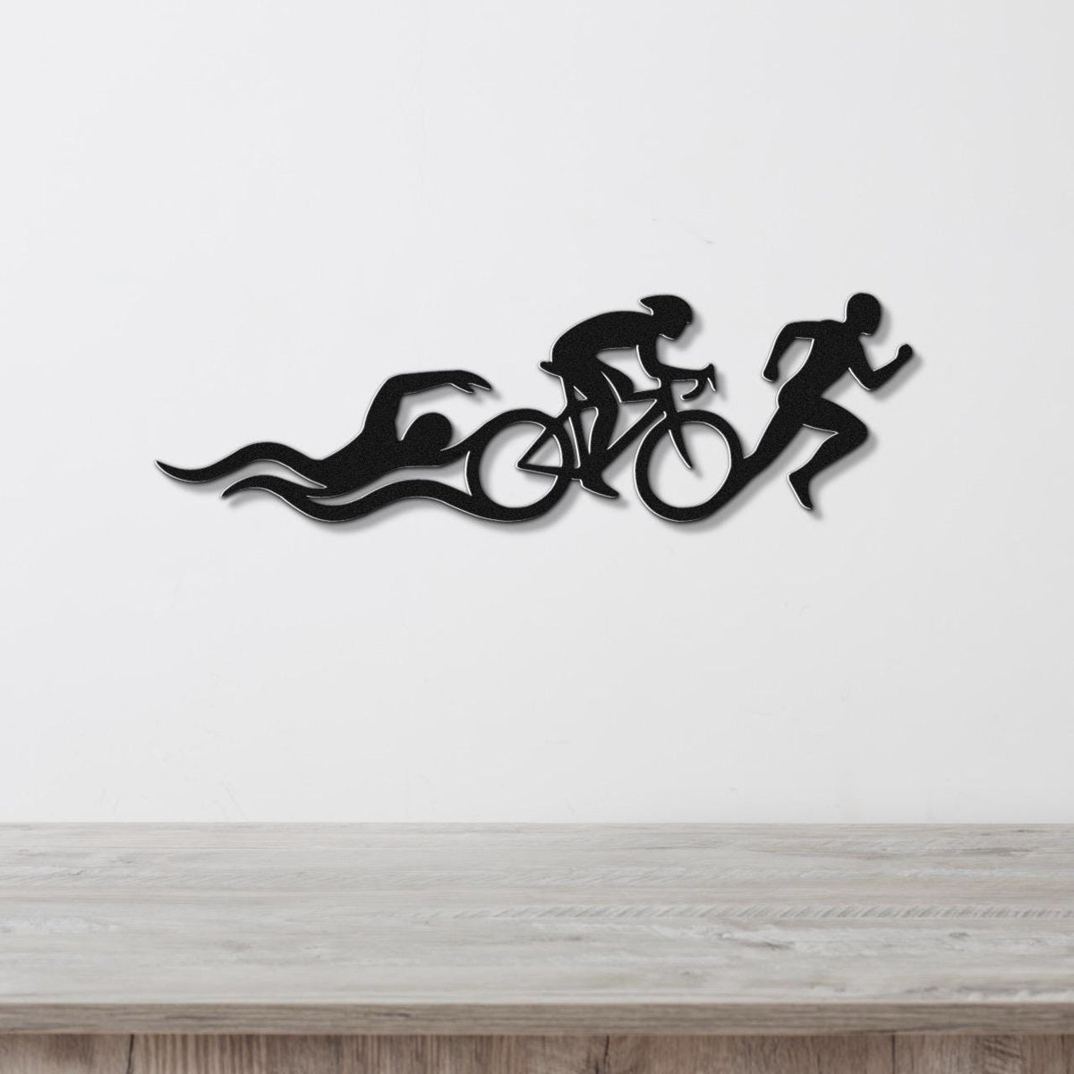 Black metal triathlon wall art with swimmer, cyclist, and runner silhouettes and motion lines, mounted on a white wall above a wooden counter.