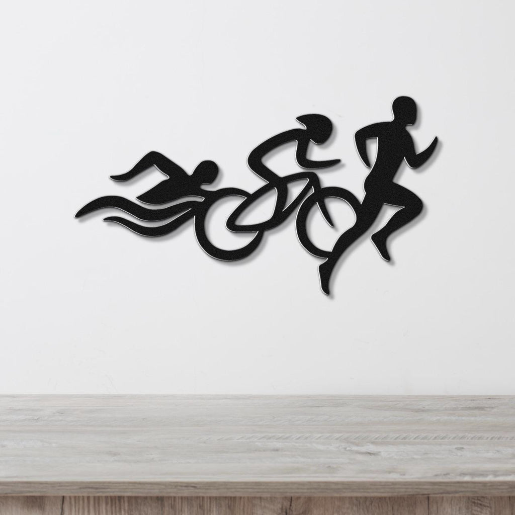 Black metal wall art featuring a triathlon silhouette of swim, bike, and run figures in motion, mounted on a white wall above a wooden surface.