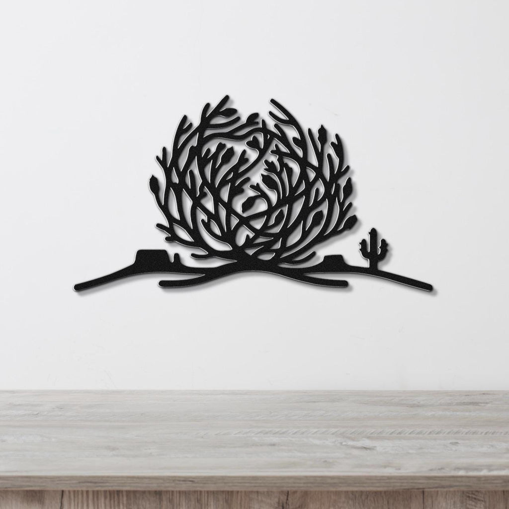Black metal wall art cutout of a tumbleweed and cactus on a desert landscape, mounted on a white wall above a wooden surface.