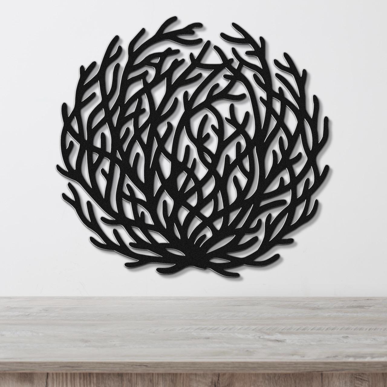 Black metal wall art in a circular, branching silhouette resembling coral or tumbleweed, mounted on a white wall above a light wood surface.