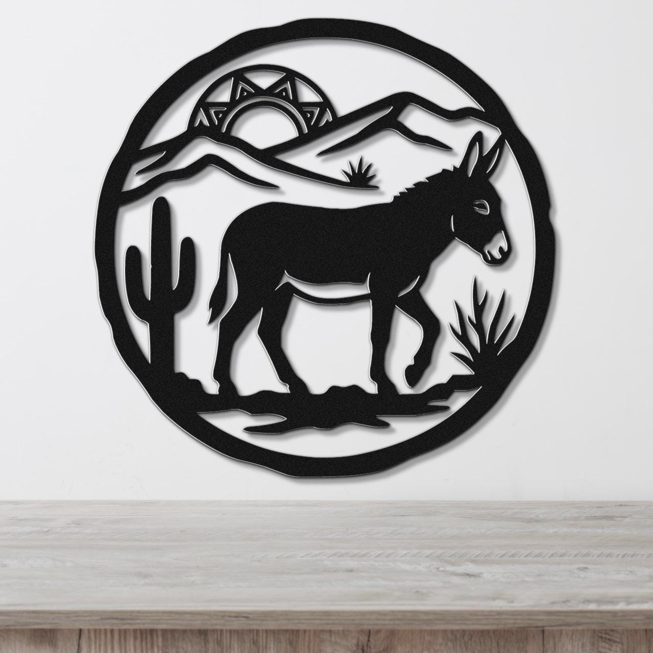 Black metal wall art featuring a burro silhouette in a desert scene with cactus, mountains, and a sun cutout, mounted on a white wall above a wooden surface.