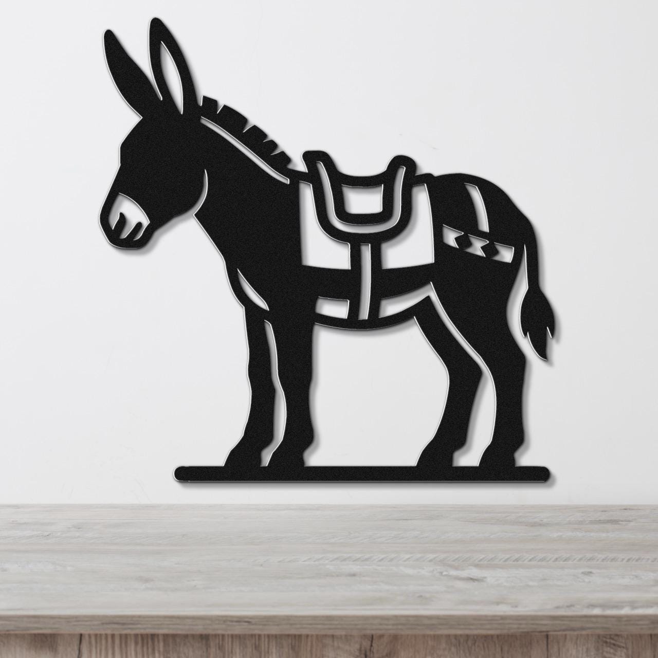 Black metal burro wall art with saddle and bridle cutouts, mounted on a white wall above a wooden surface.