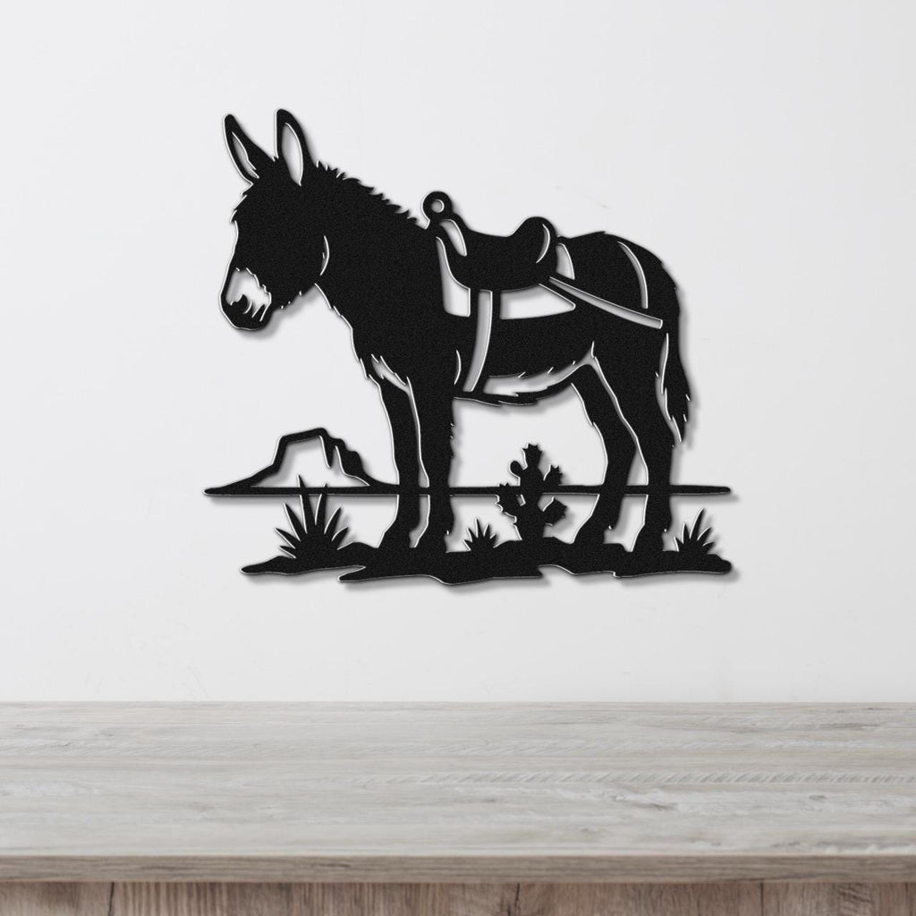 Black metal wall art silhouette of a burro with a saddle, standing on a desert landscape with cacti and a rock, mounted on a white wall above a wooden surface.