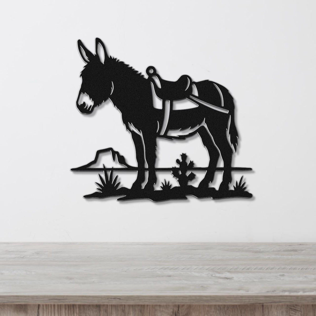 Black metal wall art silhouette of a burro with a saddle, standing on a desert landscape with cacti and a rock, mounted on a white wall above a wooden surface.