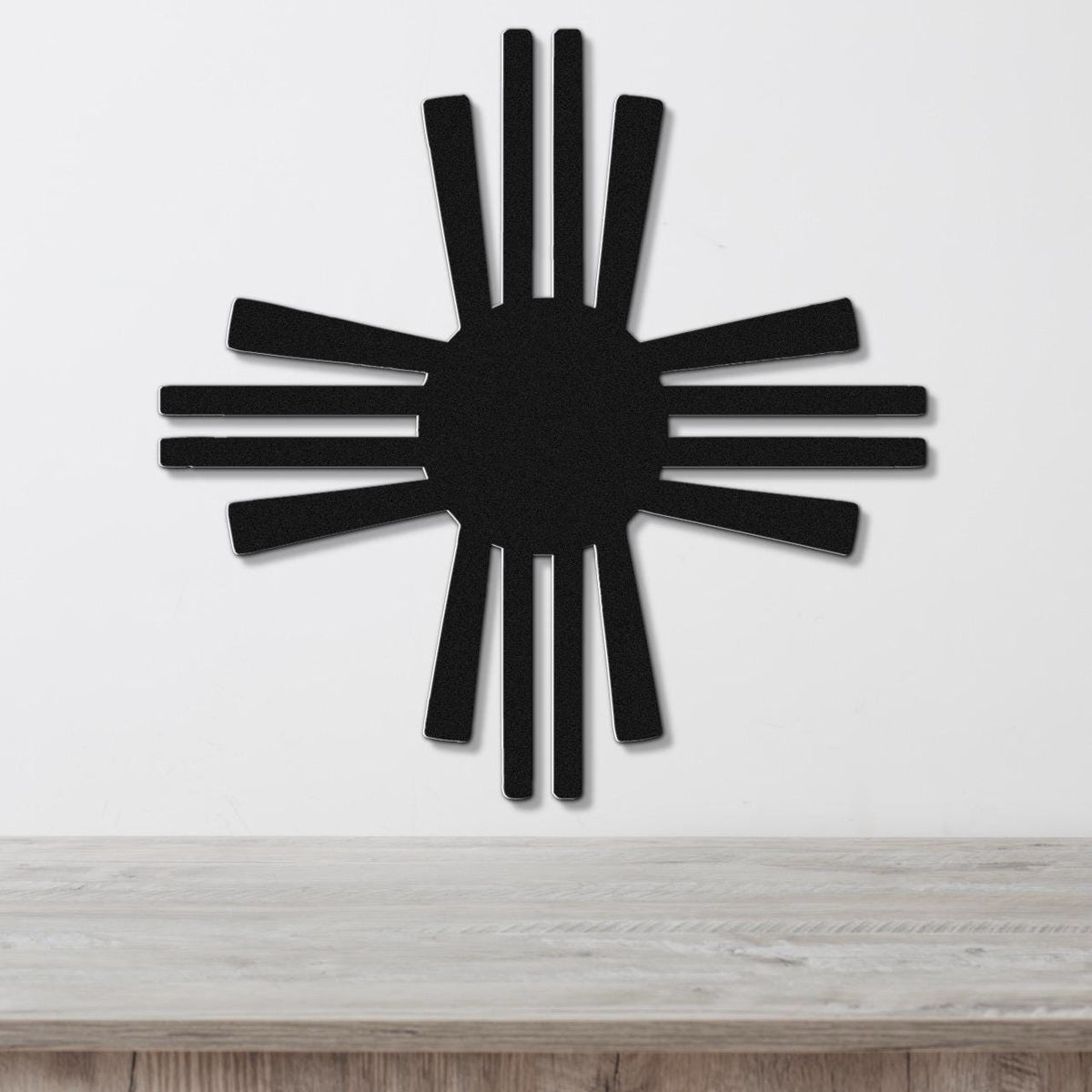 Black metal Zia Sun symbol wall art with radiating rays, mounted on a white wall above a light wood surface.