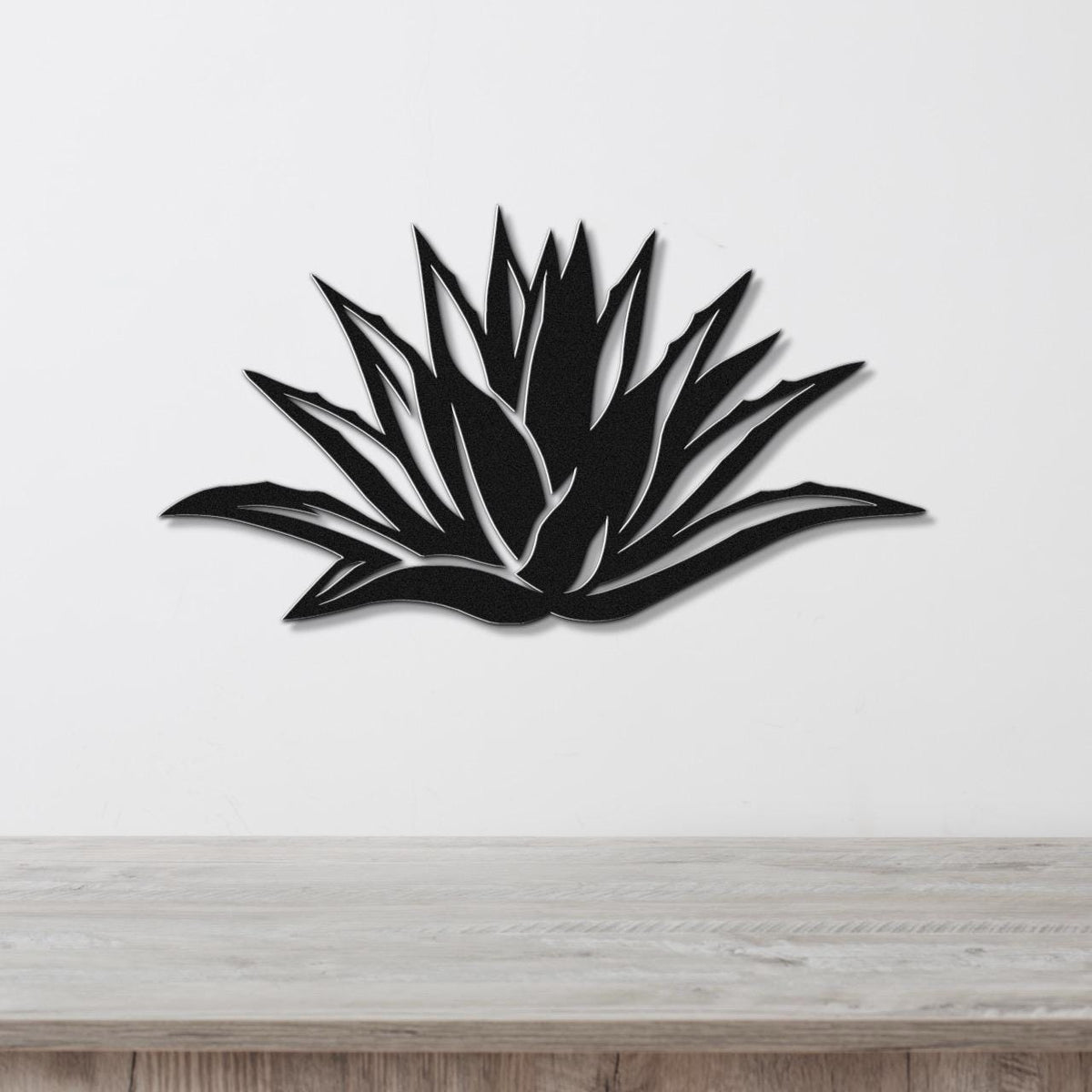 Black metal agave wall art with sharp, pointed leaf cutouts, mounted on a white wall above a light wood surface.