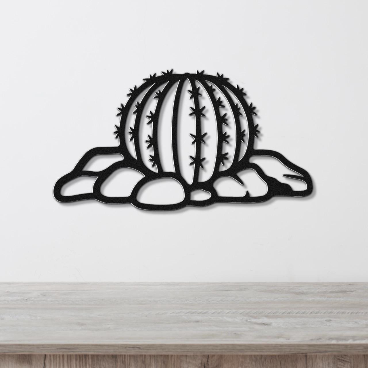 Black metal barrel cactus wall art with spines and rocks cutout, mounted on a white wall above a wooden counter.