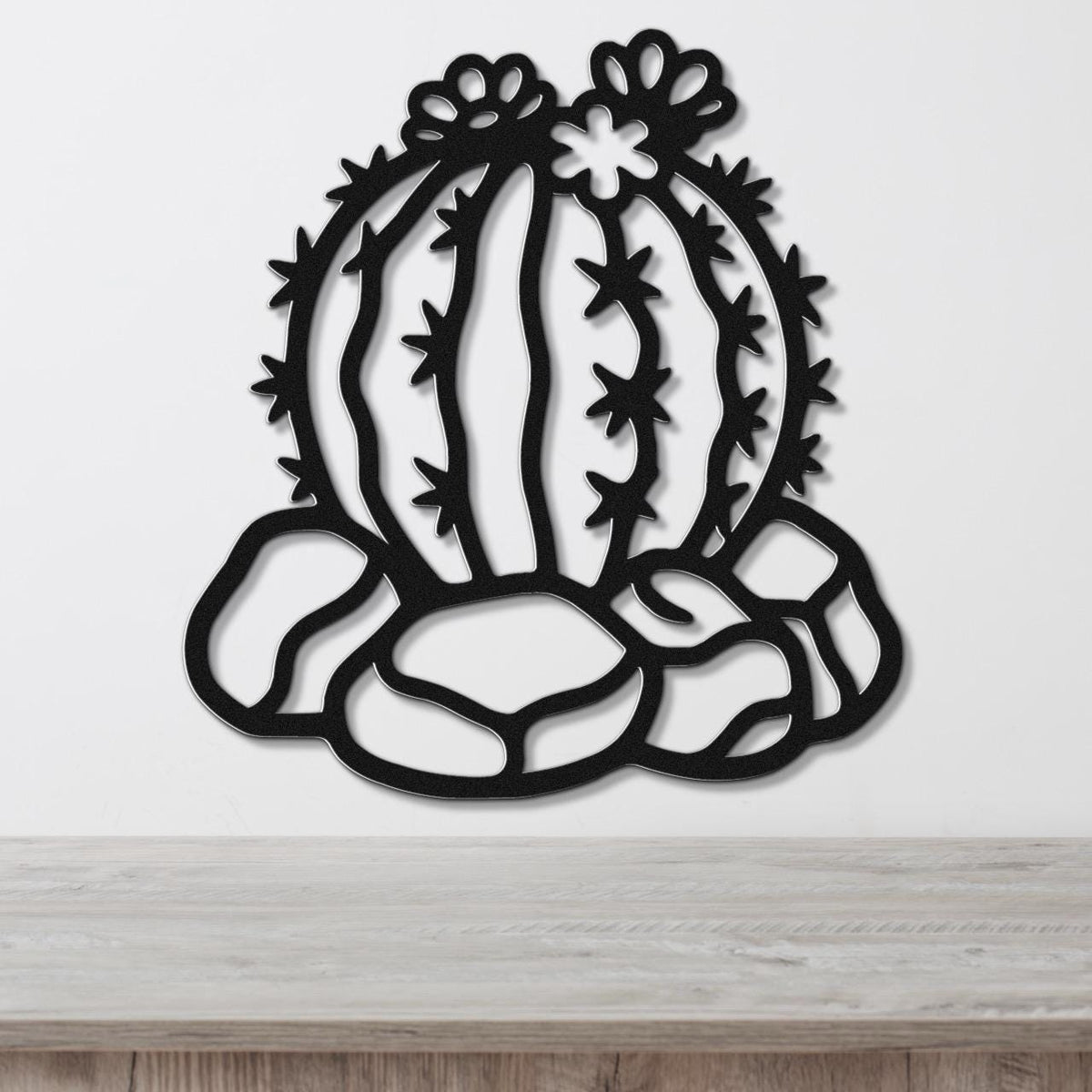 Black metal barrel cactus wall art with flower and spine cutouts, mounted on a white wall above a wooden surface.