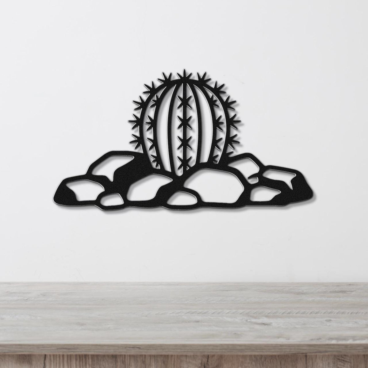Black metal barrel cactus wall art with spines and rocks cutout, mounted on a white wall above a wooden counter.