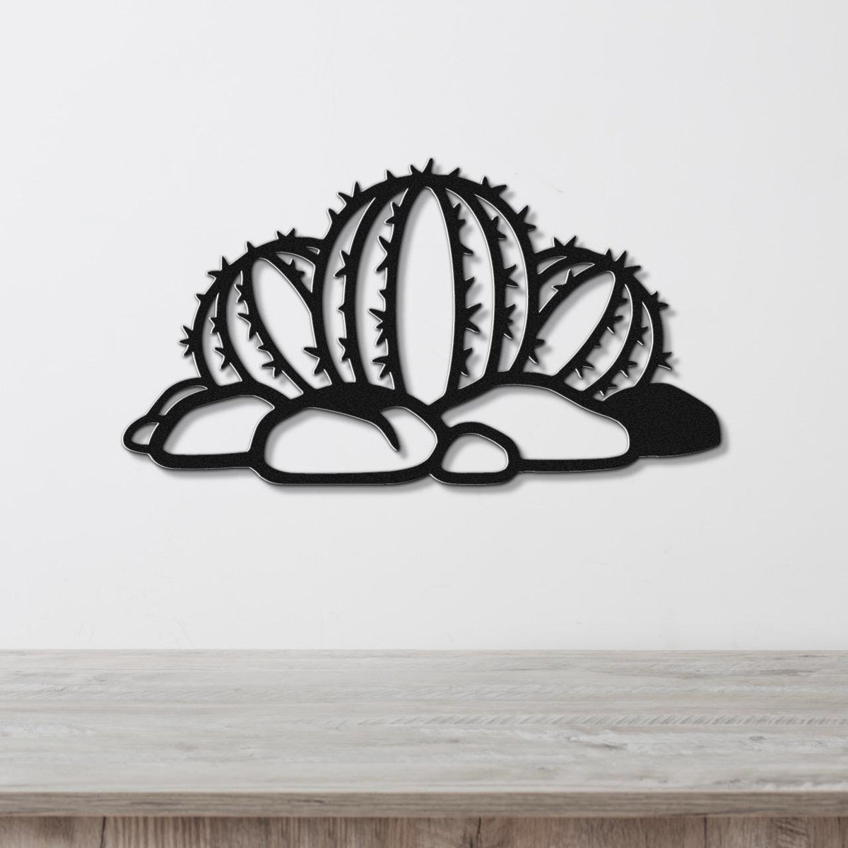 Black metal barrel cactus wall art with spines and cutout details, mounted on a white wall above a wooden surface.