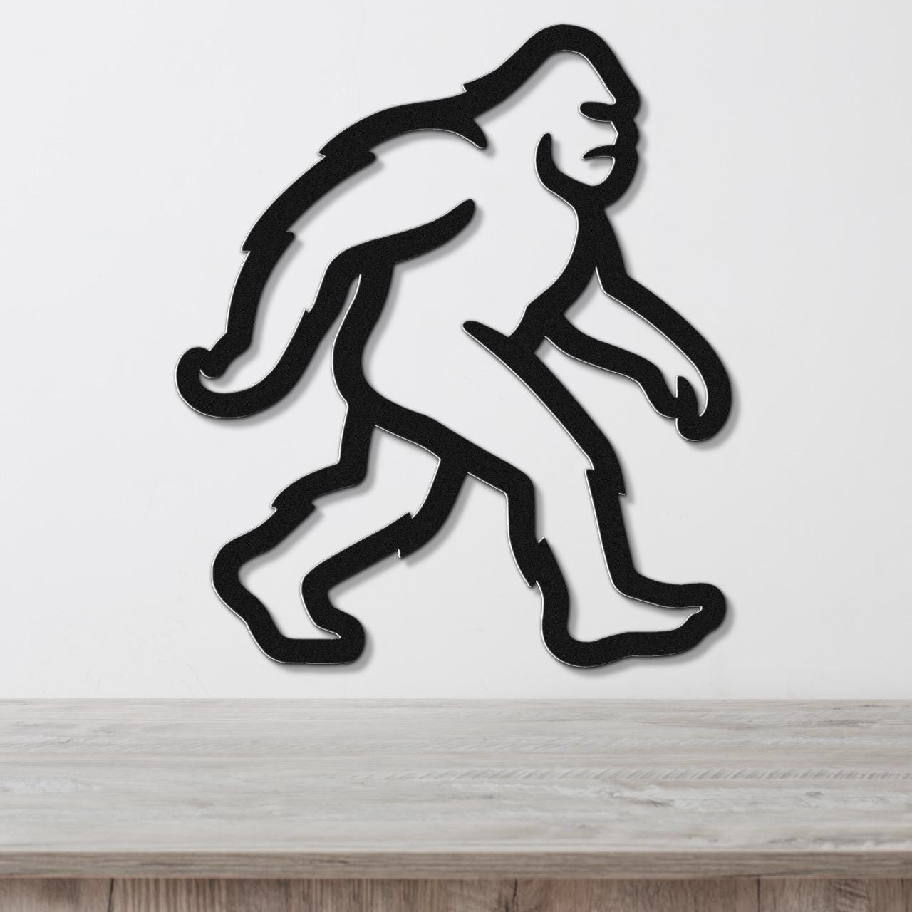 Black metal wall art cutout of a Bigfoot figure in a walking pose, mounted on a white wall above a wooden surface.