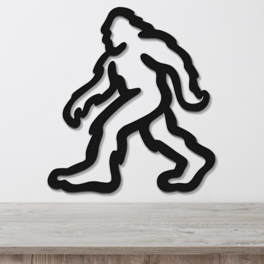 Black metal wall art featuring a minimalist Bigfoot silhouette with internal cutout details, mounted on a white wall above a light wood surface.