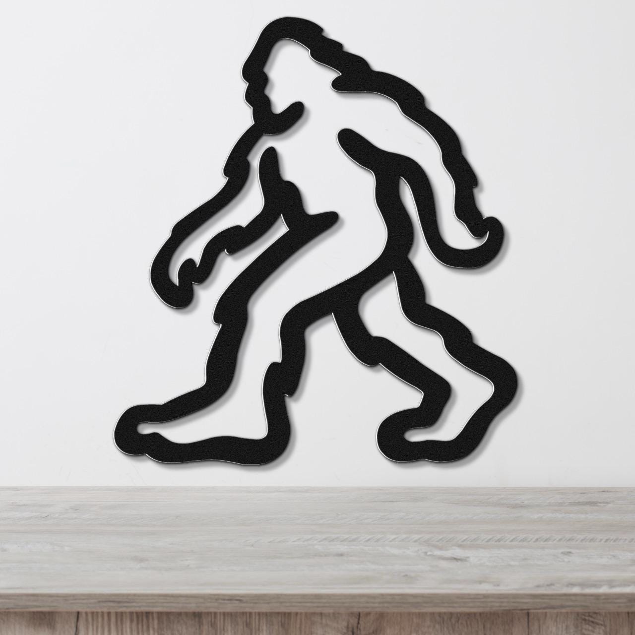Black metal wall art featuring a minimalist Bigfoot silhouette with internal cutout details, mounted on a white wall above a light wood surface.
