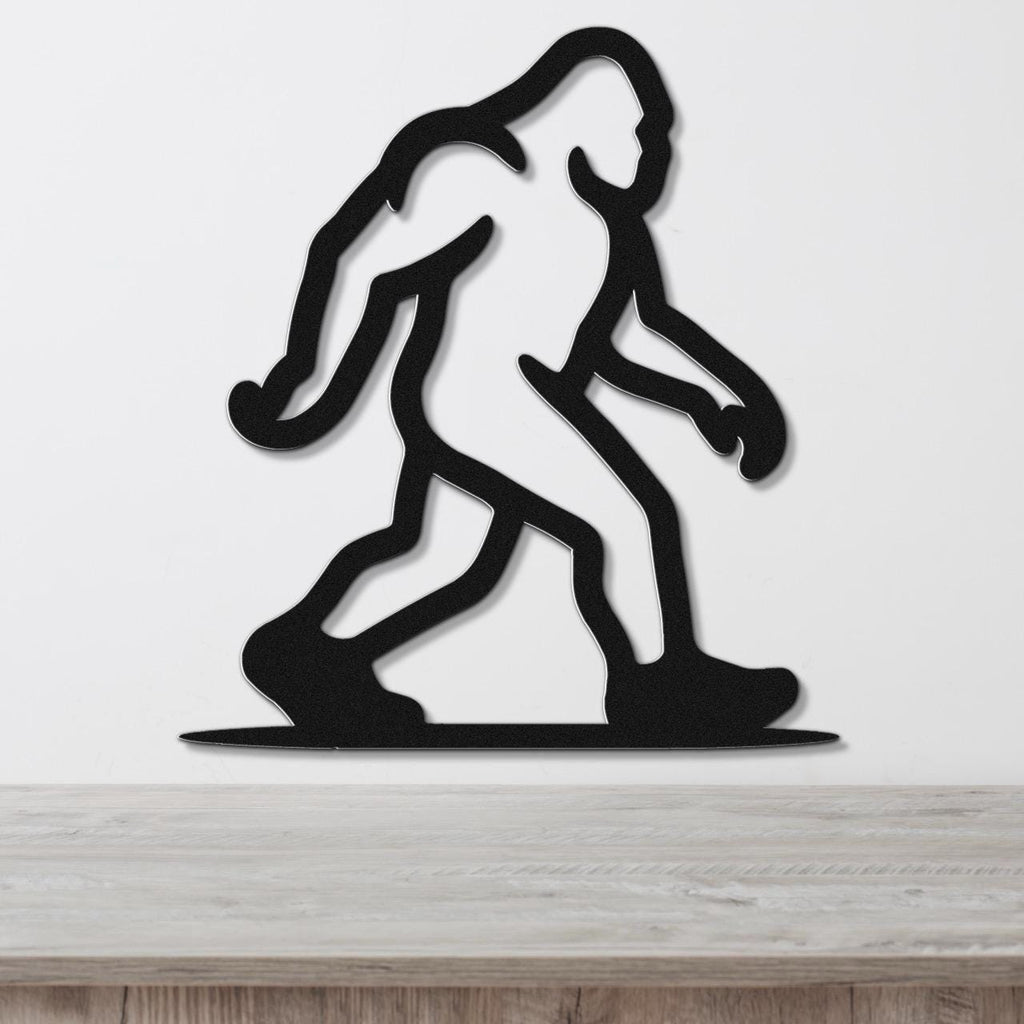 Black metal wall art silhouette of Bigfoot in a walking pose, mounted on a white wall above a light wood surface.