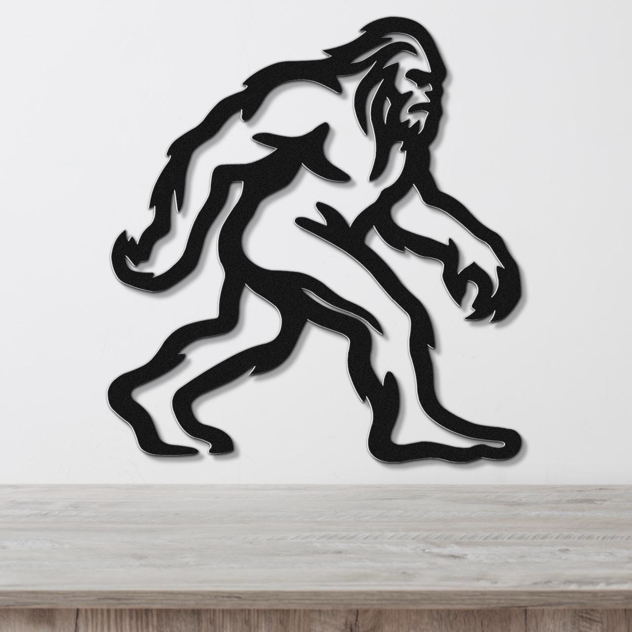 Black metal wall art featuring a rugged Bigfoot silhouette with internal cutout details, mounted on a white wall above a wooden surface.