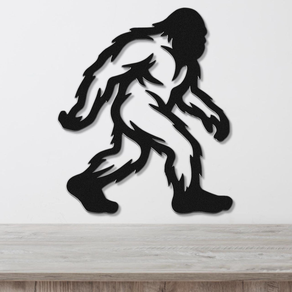 Black metal Bigfoot wall art silhouette with internal cutout details, mounted on a white wall above a wooden surface.