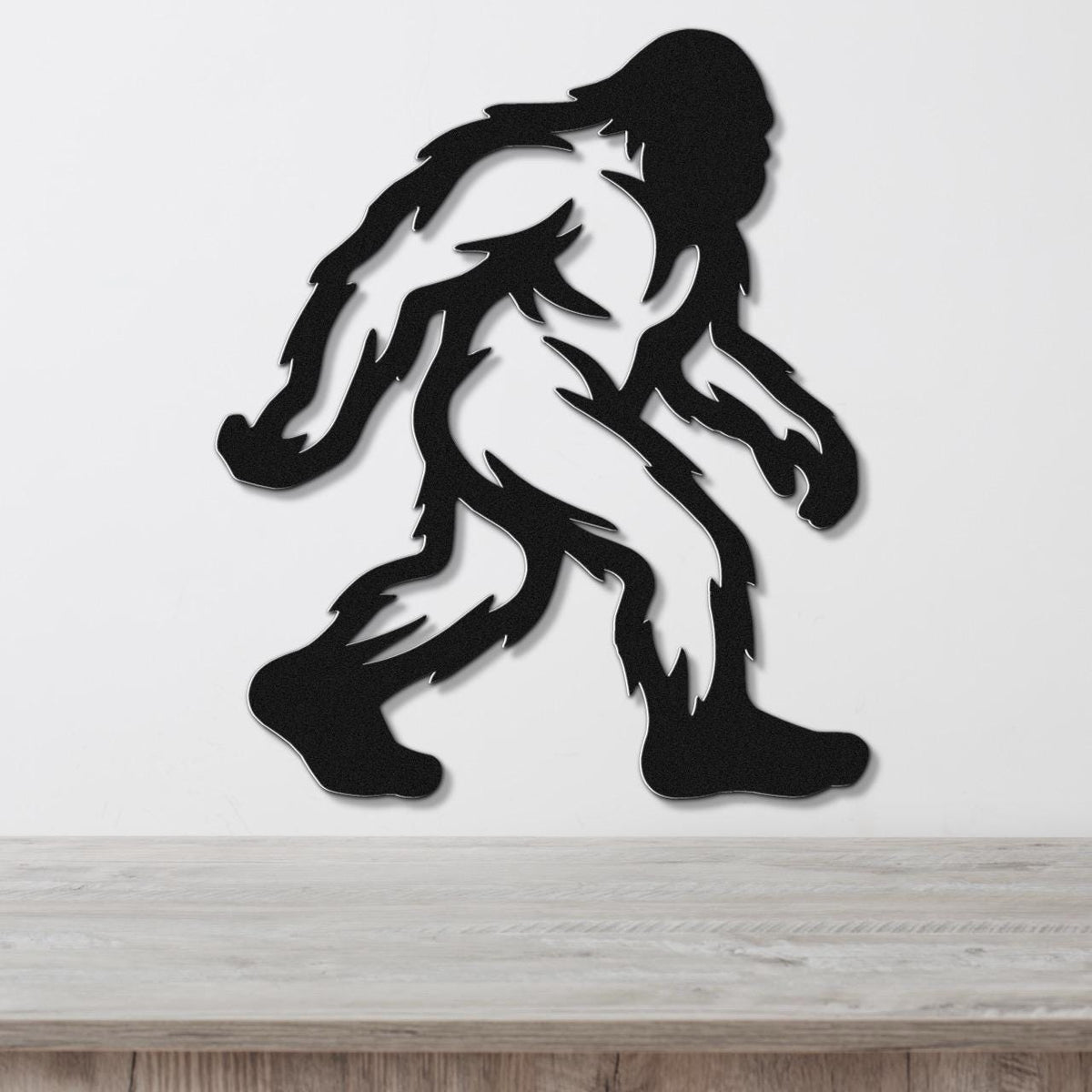 Black metal Bigfoot wall art silhouette with internal cutout details, mounted on a white wall above a wooden surface.