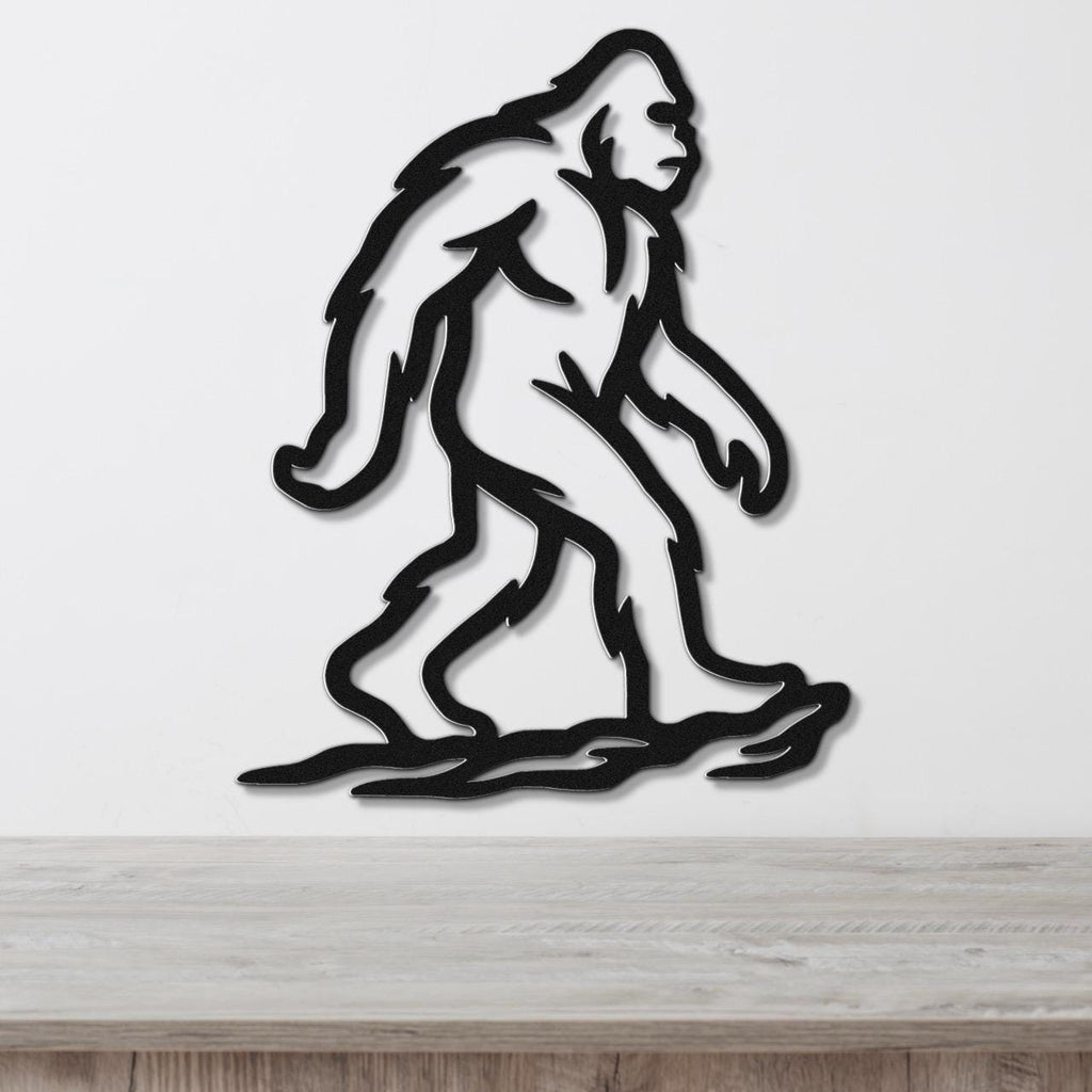 Black metal Bigfoot silhouette wall art with internal cutout details, mounted on a white wall above a light wood surface.