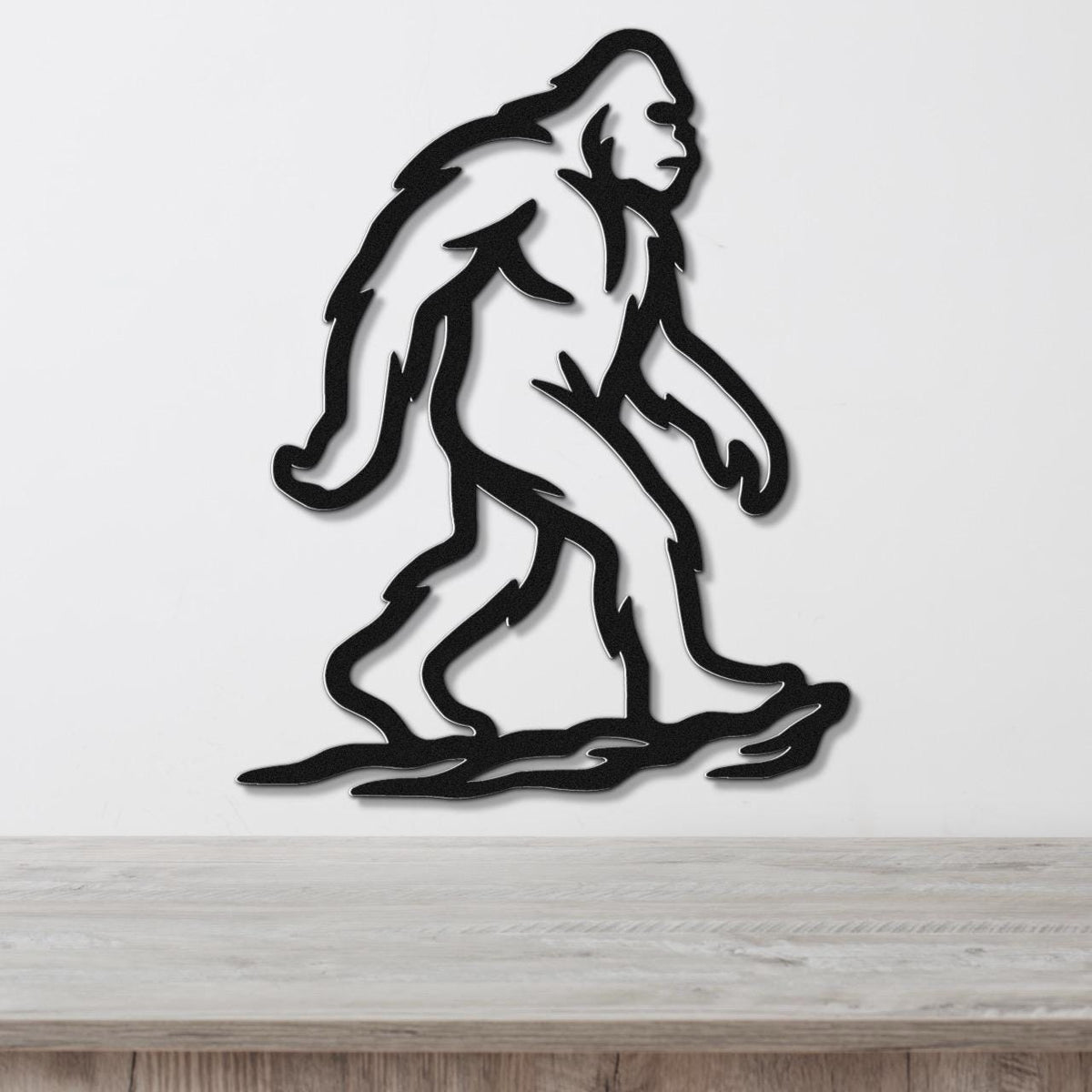 Black metal Bigfoot silhouette wall art with internal cutout details, mounted on a white wall above a light wood surface.