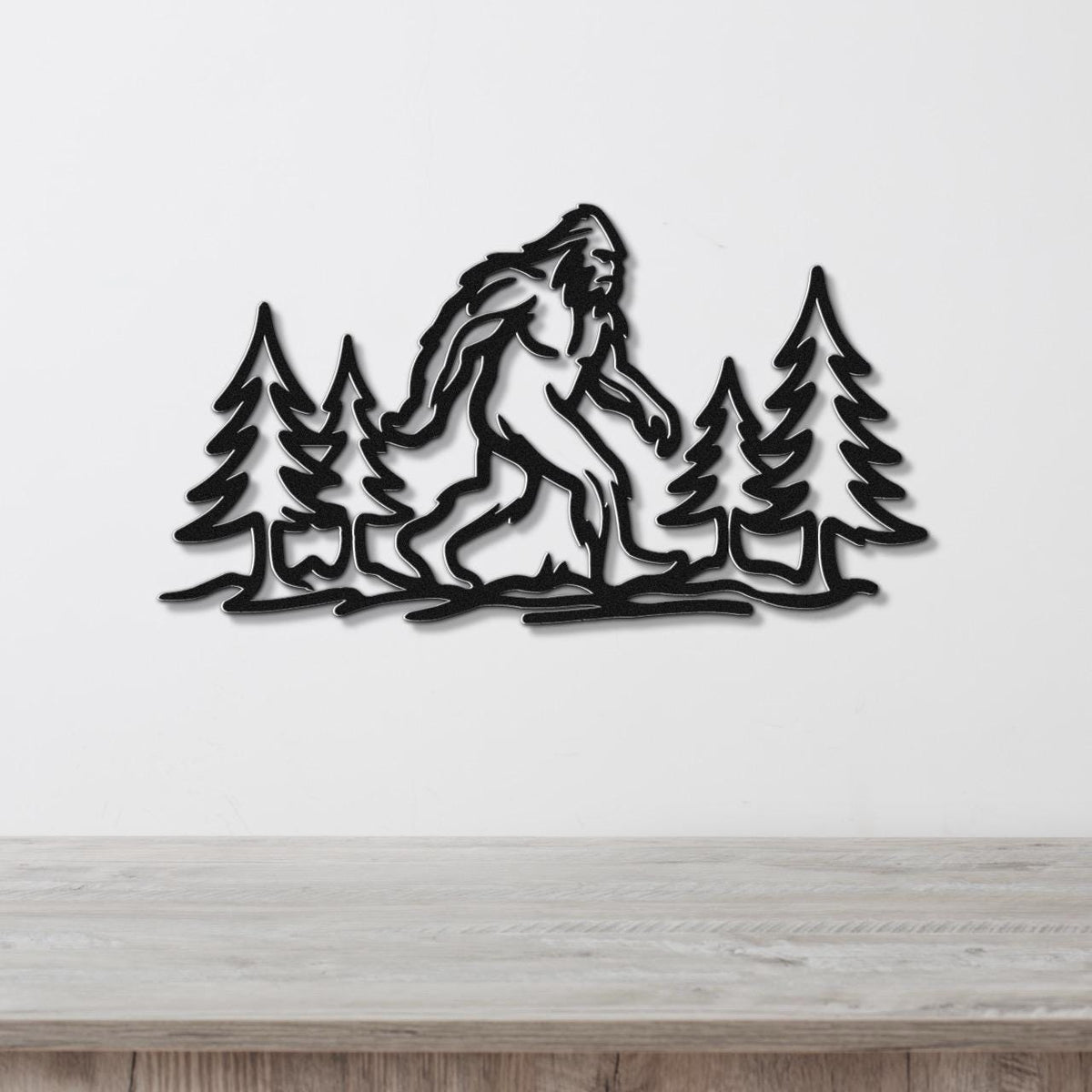 Black metal wall art featuring a Bigfoot silhouette walking through pine trees, mounted on a white wall above a wooden counter.