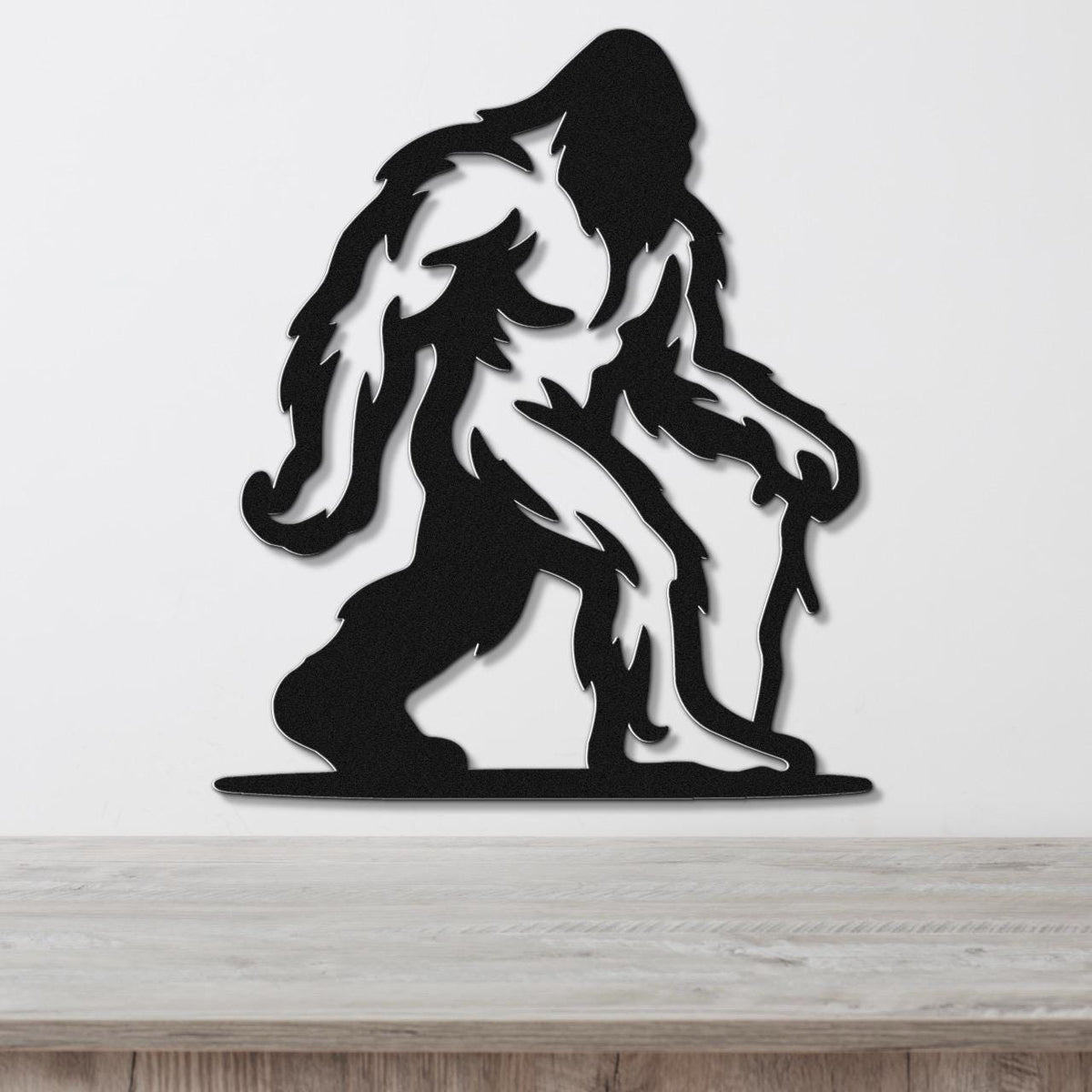 Black metal wall art silhouette of Bigfoot with detailed fur cutouts, mounted on a white wall above a wooden surface.