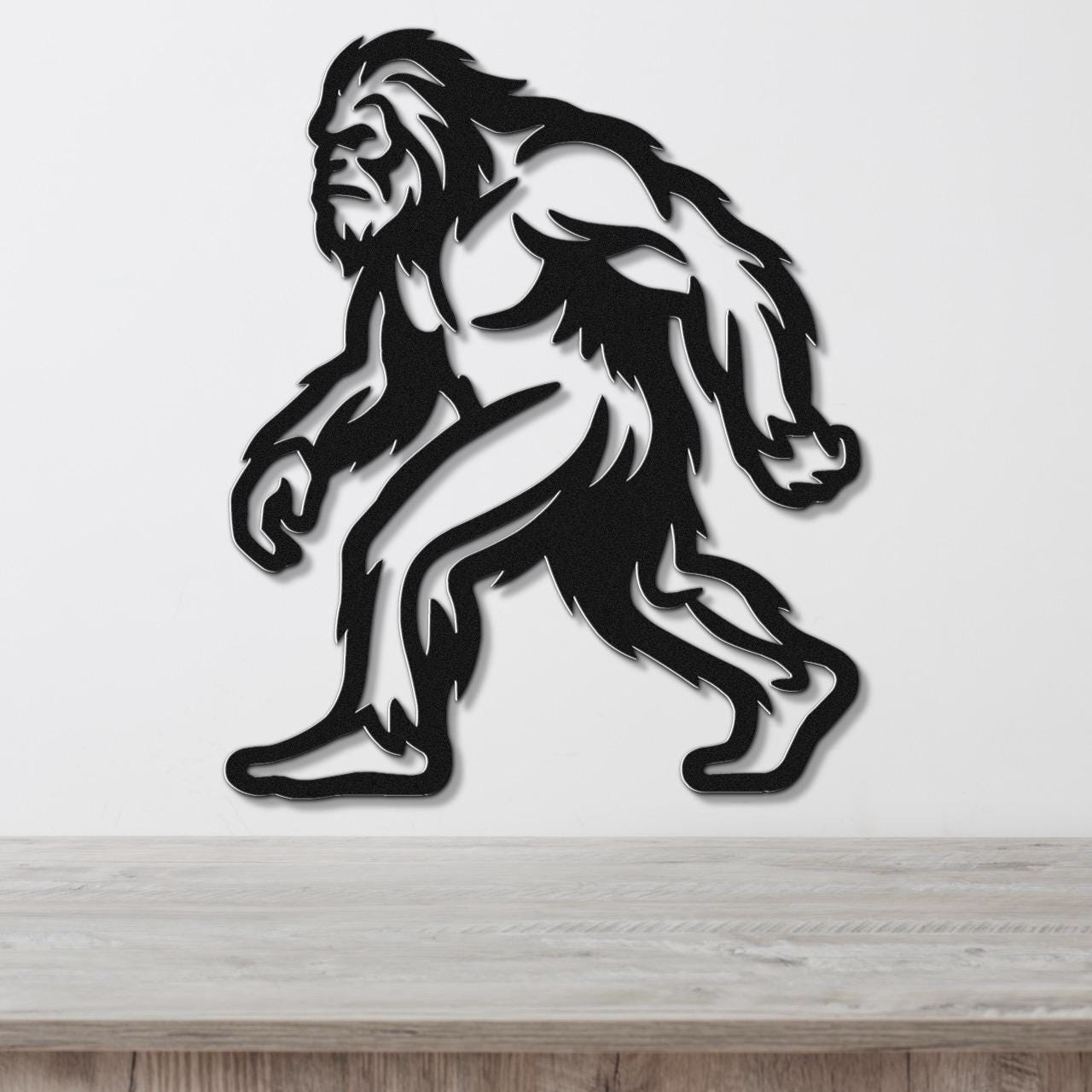 Black metal wall art silhouette of Bigfoot with detailed fur cutouts, mounted on a white wall above a wooden surface.