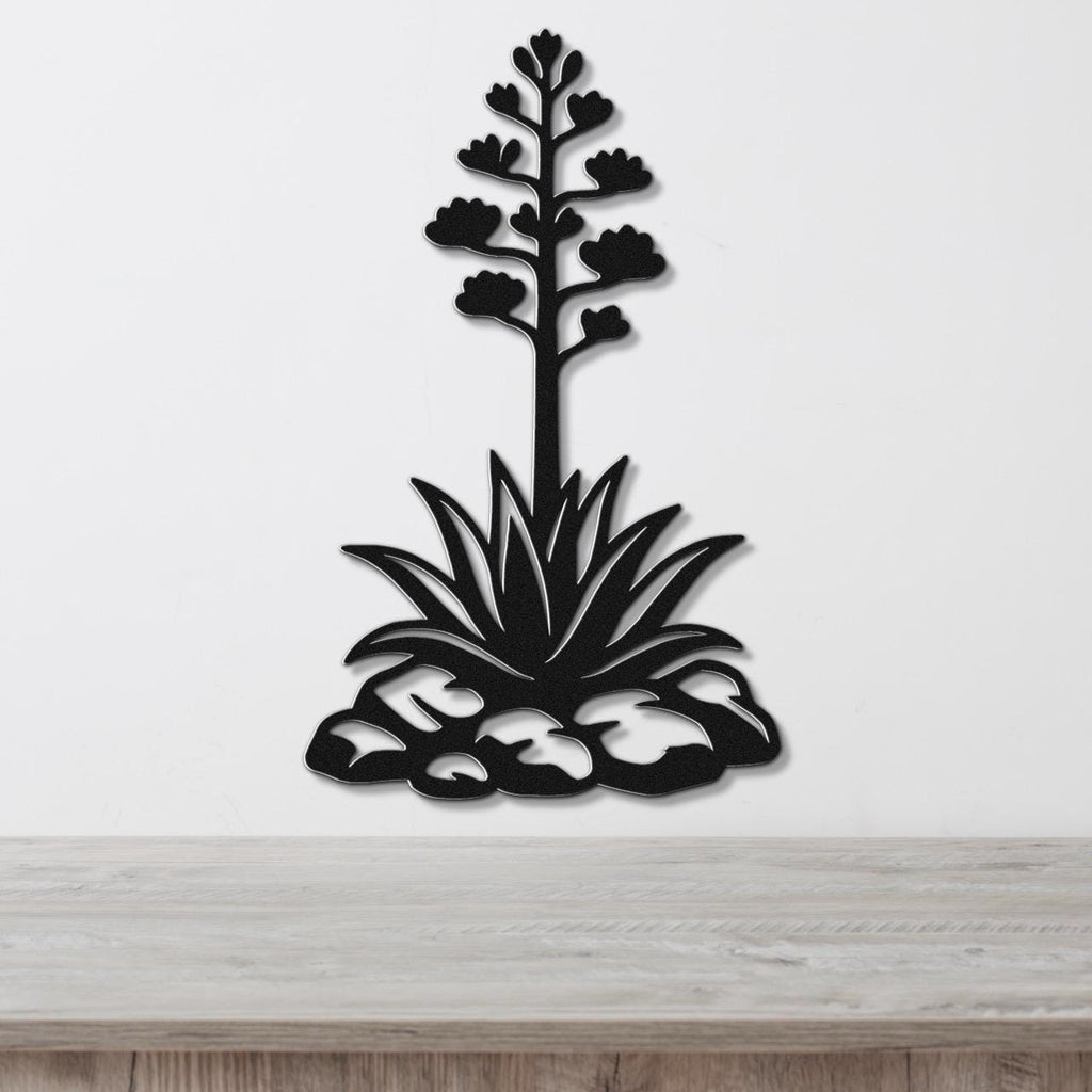 Black metal agave wall sculpture with blooming flower stalk and spiky leaves, mounted on a white wall above a wooden surface.