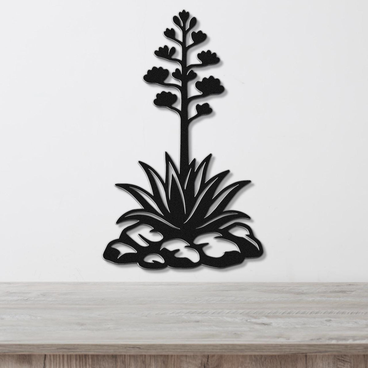 Black metal agave wall sculpture with blooming flower stalk and spiky leaves, mounted on a white wall above a wooden surface.