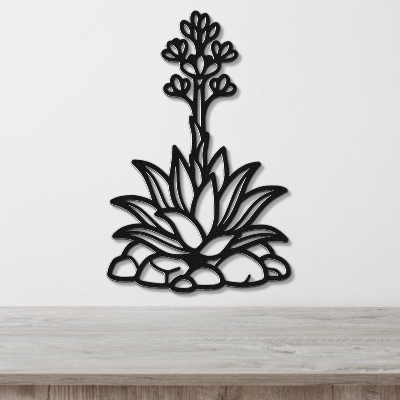 Black metal agave wall art with blooming flower cutout, mounted on a white wall above a wooden surface.