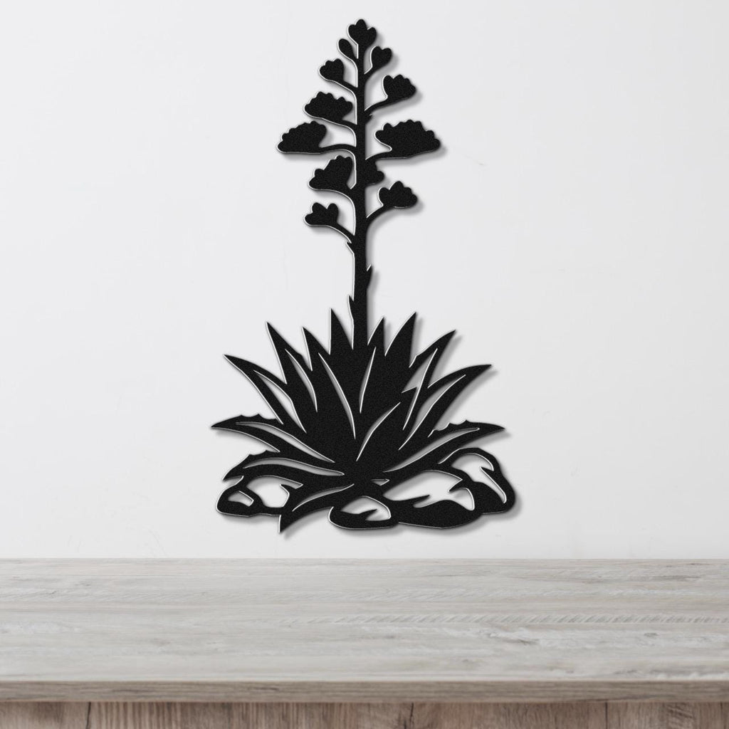 Black metal wall art silhouette of a blooming agave plant with a tall flower stalk and spiky leaves, mounted on a white wall above a wooden surface.