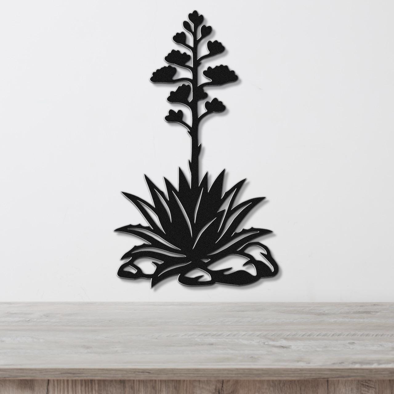 Black metal wall art silhouette of a blooming agave plant with a tall flower stalk and spiky leaves, mounted on a white wall above a wooden surface.