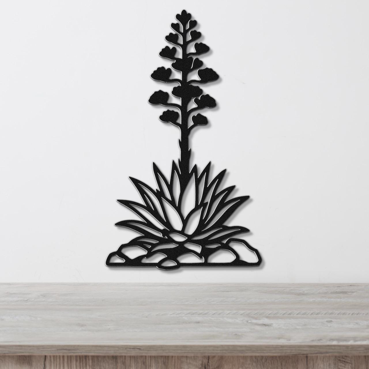 Black metal wall art in the silhouette of a blooming agave plant with cutout leaves and flower stalk, mounted on a white wall above a wooden surface.