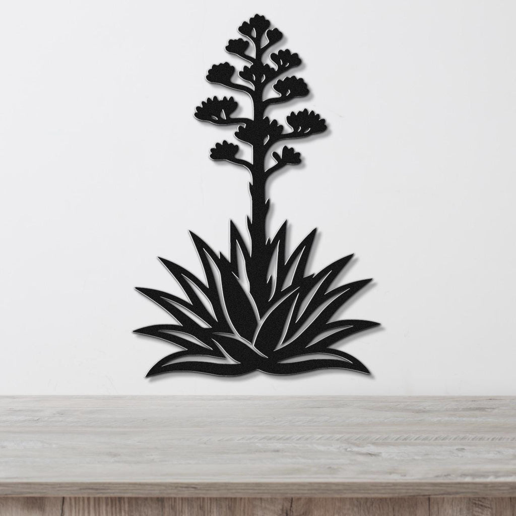 Black metal wall art in the silhouette of a blooming agave plant with cutout leaves and flower stalk, mounted on a white wall above a wooden surface.