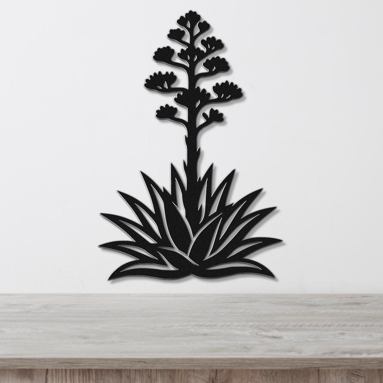 Black metal wall art in the silhouette of a blooming agave plant with cutout leaves and flower stalk, mounted on a white wall above a wooden surface.