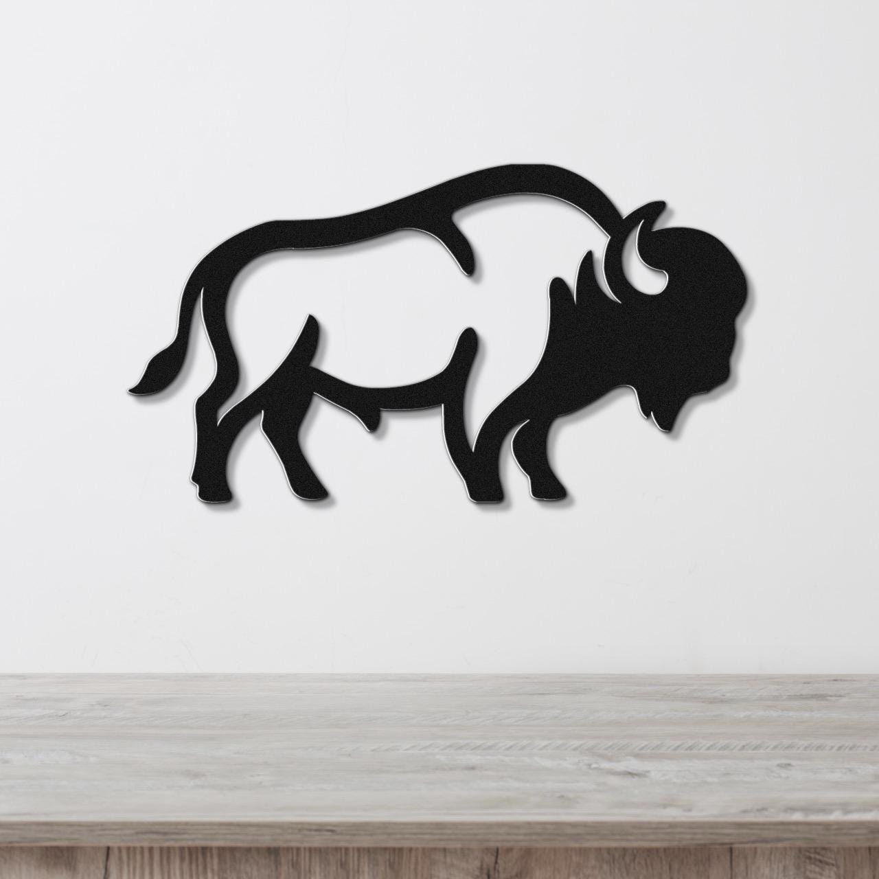 Black metal buffalo silhouette wall art with internal cutout details, mounted on a white wall above a light wood surface.