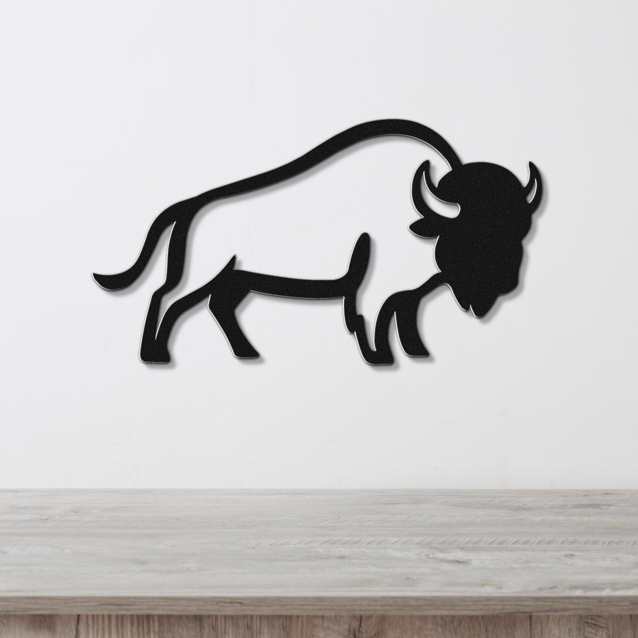 Black metal buffalo wall art silhouette with curved horns and tail, mounted on a white wall above a light wood surface.