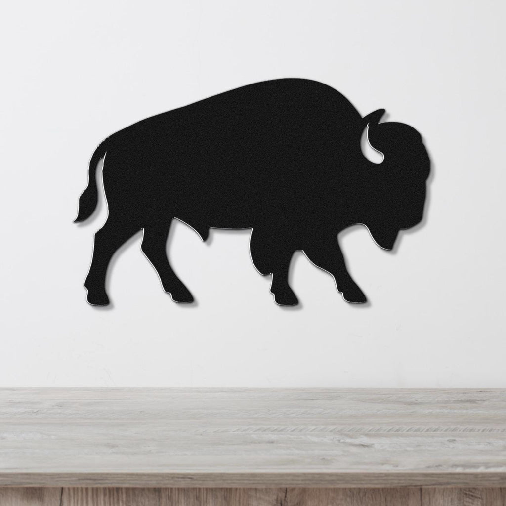 Black metal buffalo wall art silhouette with horn and tail cutouts, mounted on a white wall above a light wood surface.