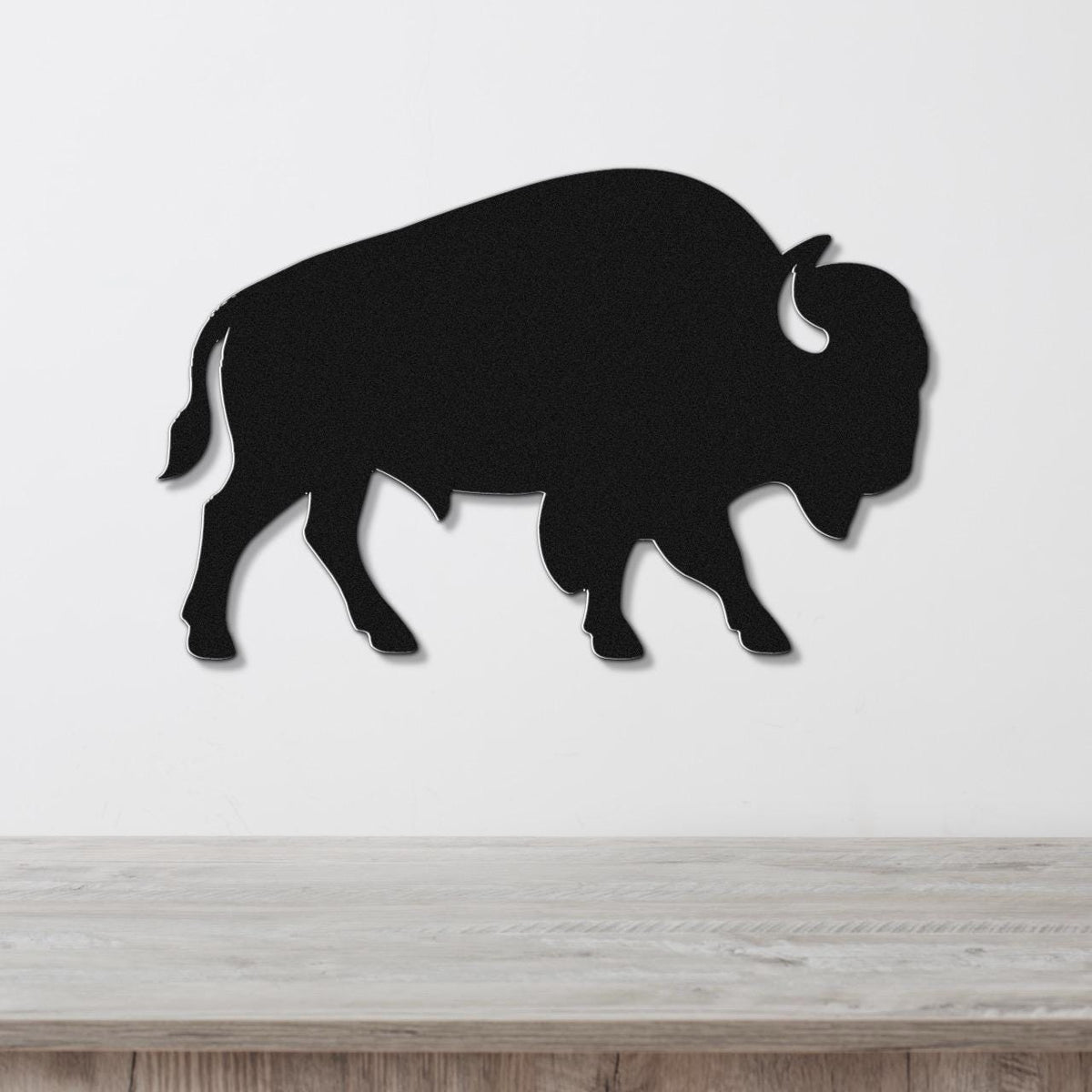Black metal buffalo wall art silhouette with horn and tail cutouts, mounted on a white wall above a light wood surface.