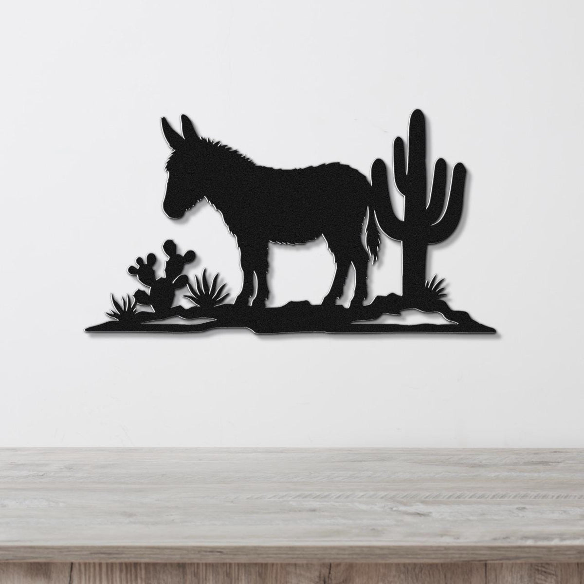 Black metal wall art silhouette of a burro and cacti, mounted on a white wall above a wooden surface.