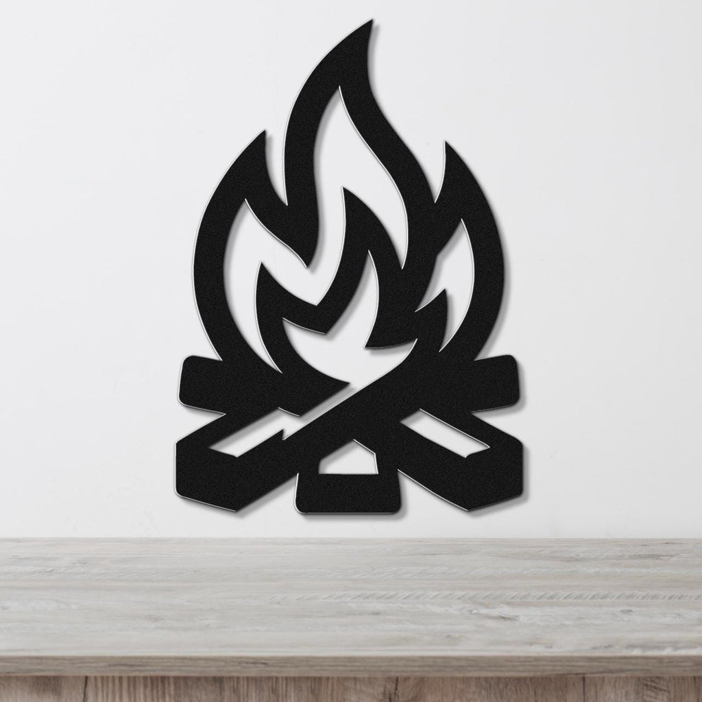 Black metal wall art in a campfire silhouette with flame and log cutouts, mounted on a white wall above a wooden surface.