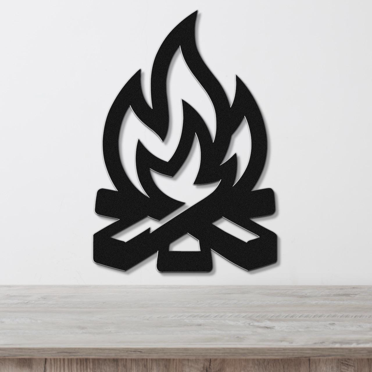 Black metal wall art in a campfire silhouette with flame and log cutouts, mounted on a white wall above a wooden surface.