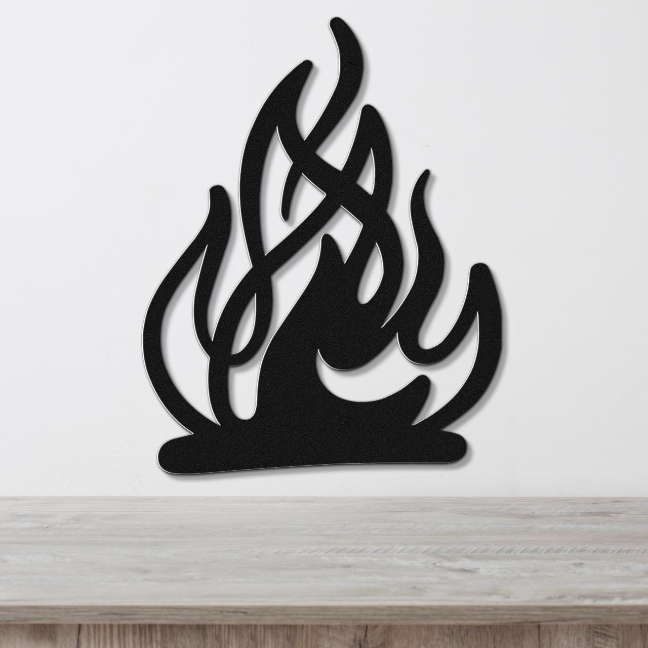 Black metal wall art in a flame silhouette design with internal cutout details, mounted on a white wall above a light wood surface.