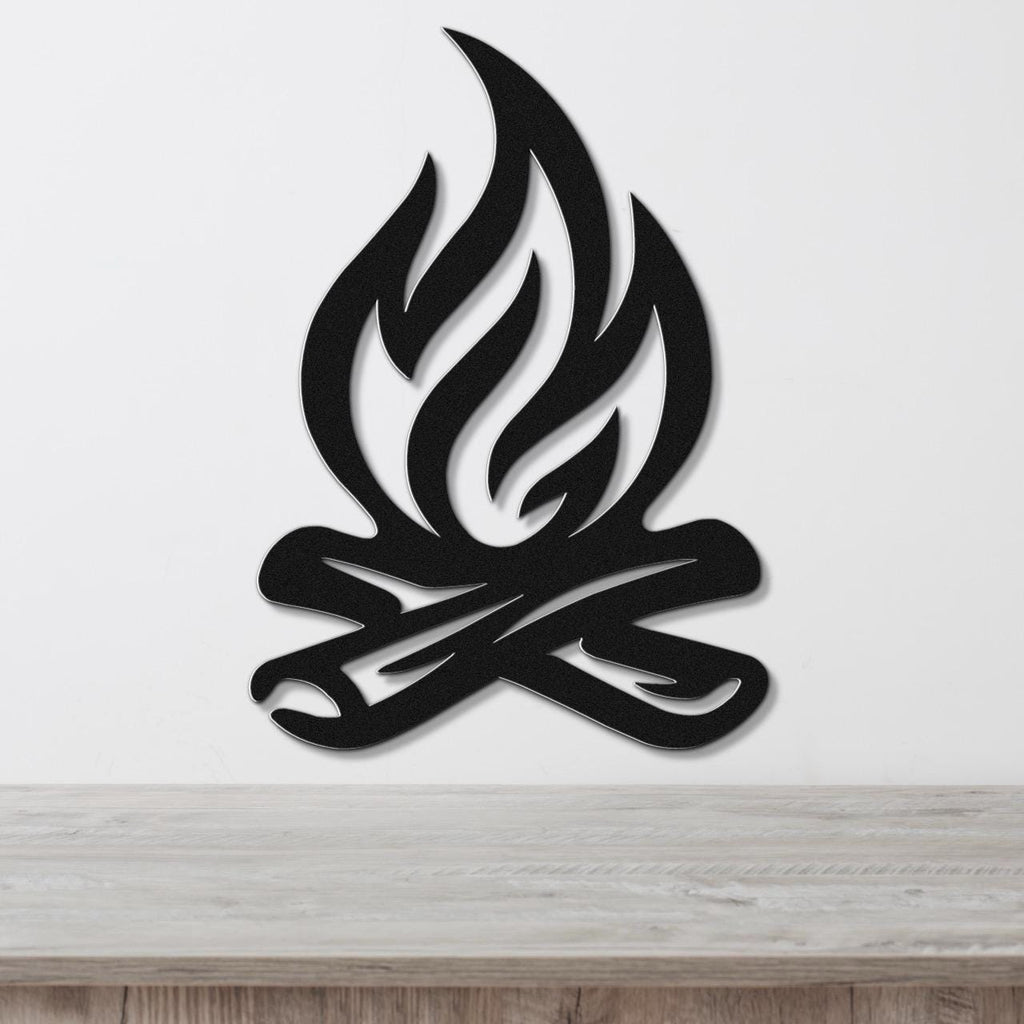 Black metal wall art in a campfire silhouette design with flame and log cutouts, mounted on a white wall above a wooden surface.