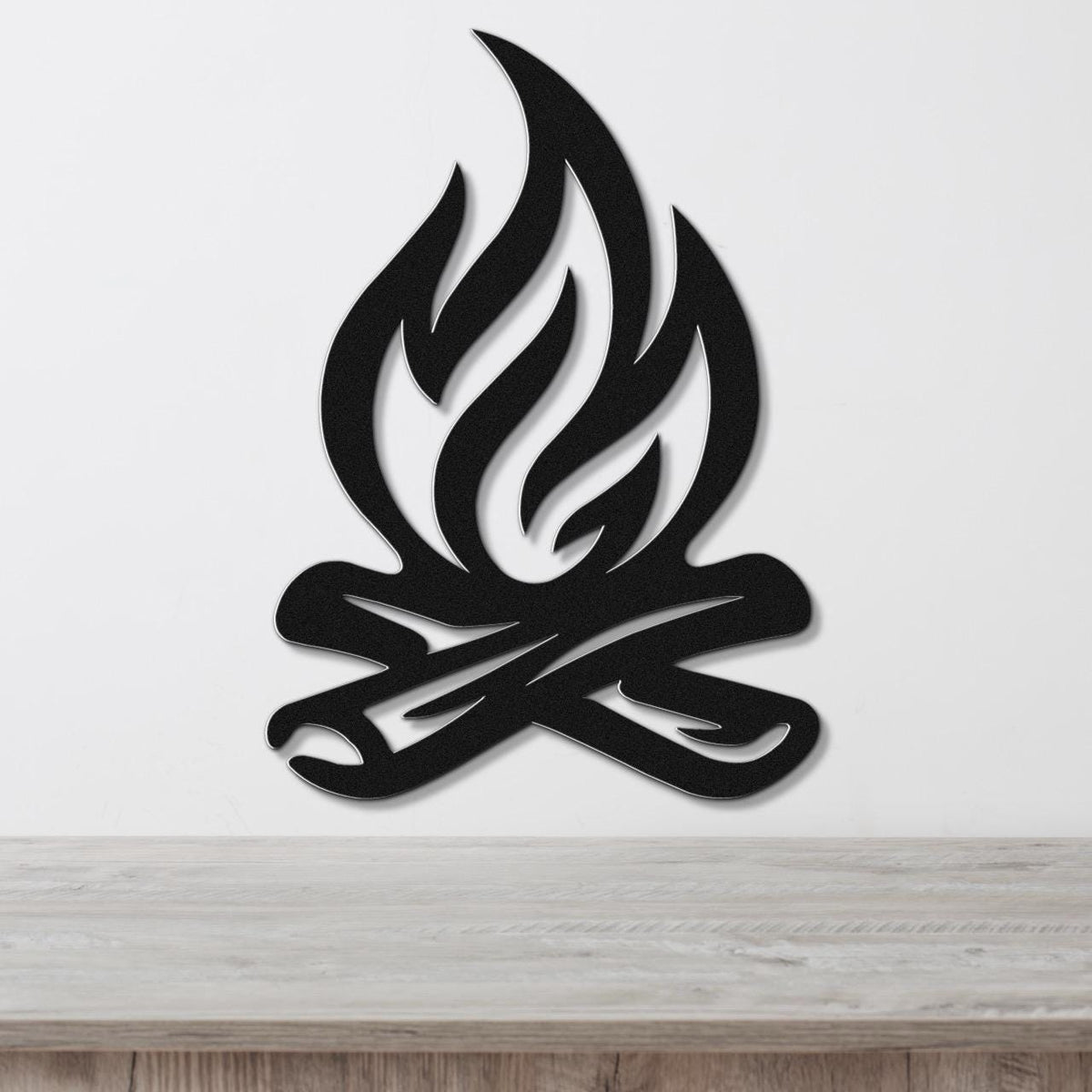 Black metal wall art in a campfire silhouette design with flame and log cutouts, mounted on a white wall above a wooden surface.