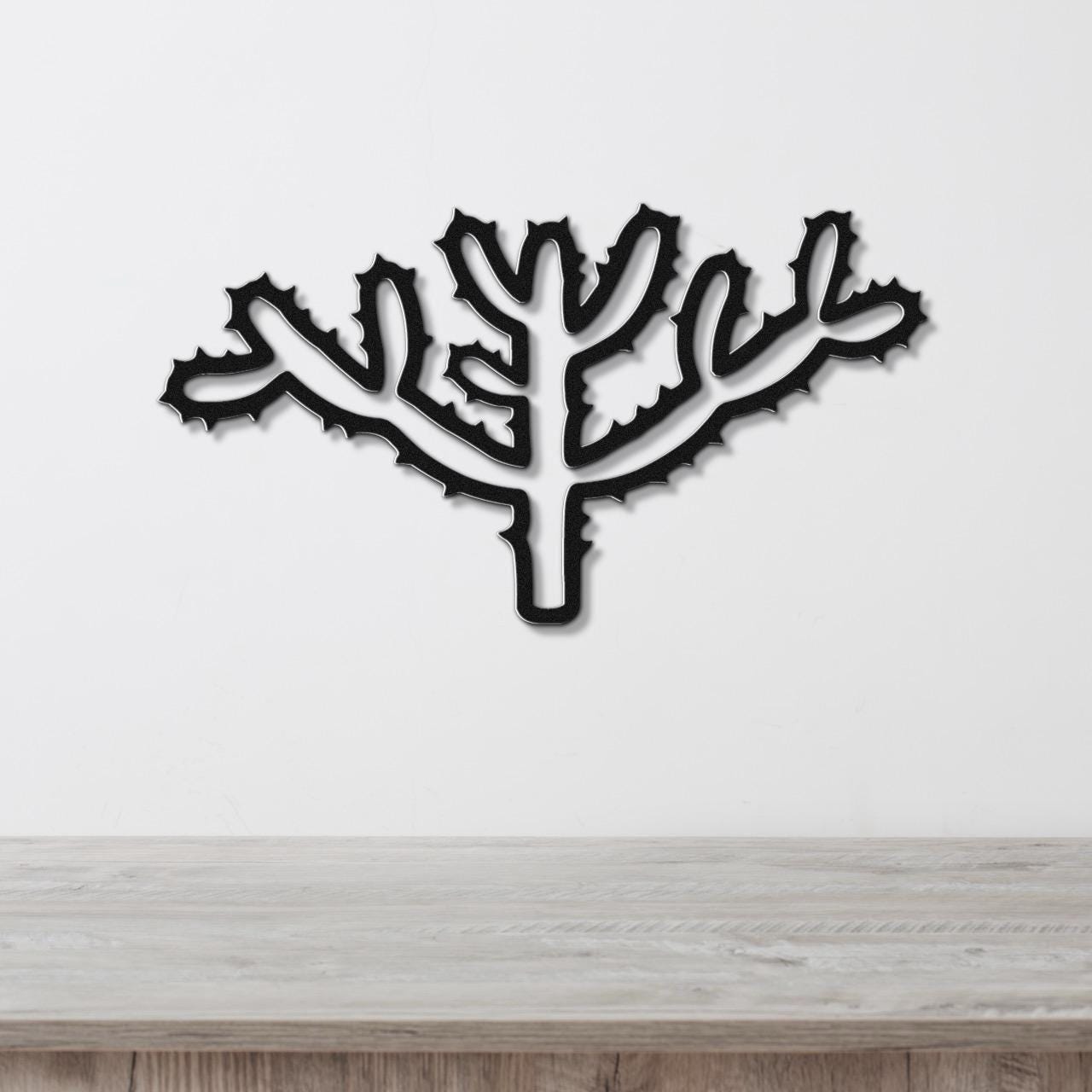 Black metal wall art cutout of a cholla cactus with spiny branches, mounted on a white wall above a light wood surface.